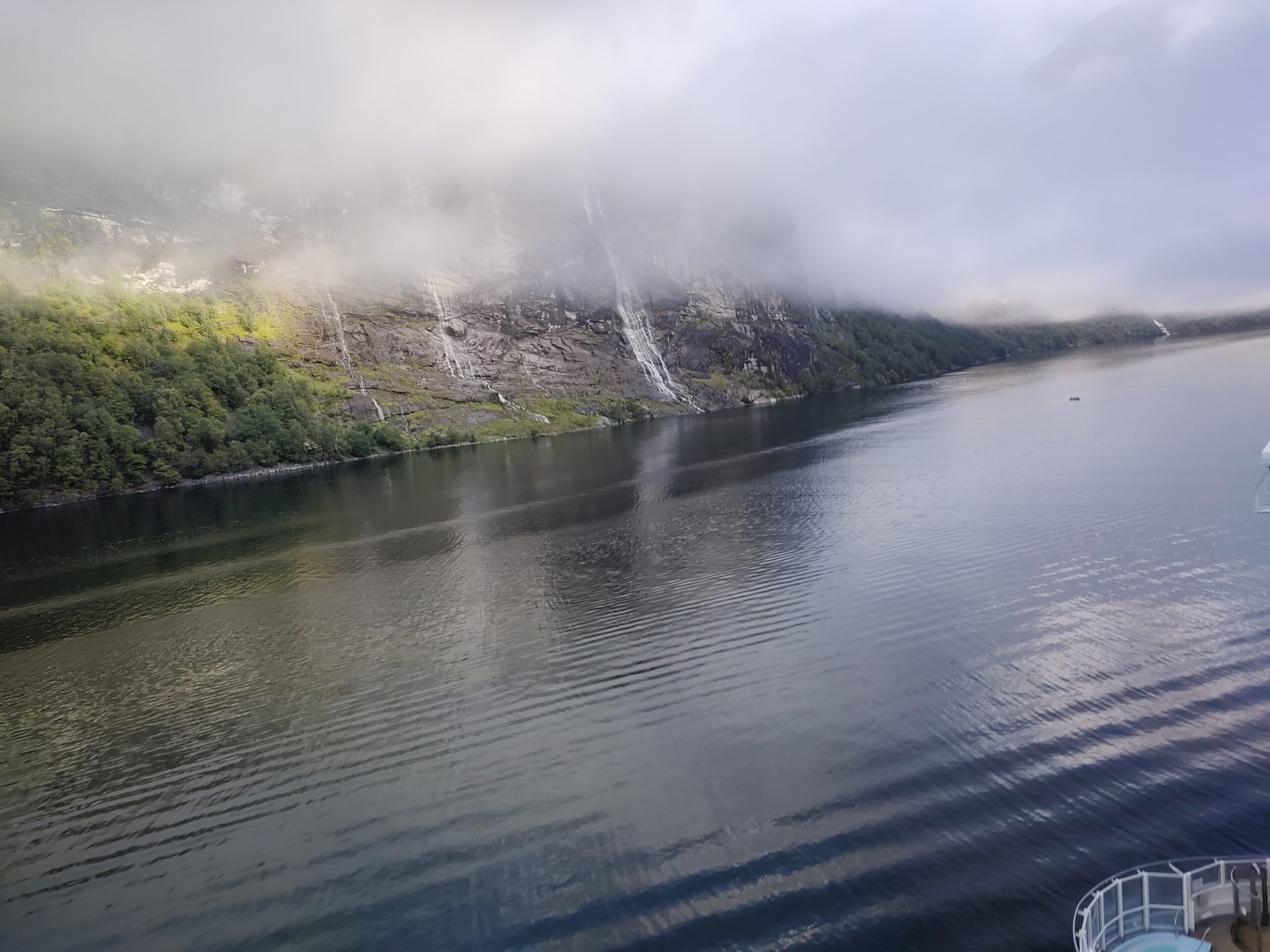 Blick vom Kreuzfahrtschiff auf den Geirangerfjord mit steilen Felswänden, Wasserfällen und tief hängenden Wolken.